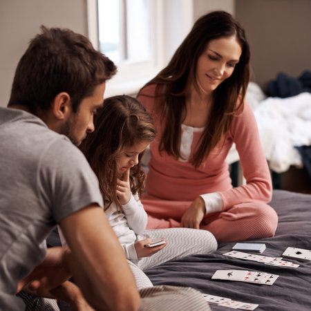 Parents, girl and playing cards to relax for game with bonding, learning and fun with strategy in home bedroom. Father, mother and daughter on bed, maths and teaching with connection in family houseの写真素材