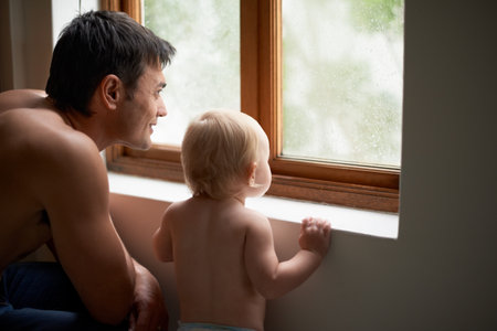 Rain, window and father with baby in a house for bonding, playing or having fun in their home together. Water, glass or dad with curious kid watching storm, weather or raindrops for learning or gamesの写真素材