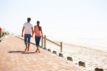Man, woman and holding hands for walk on beach, back view of romantic date and commitment with trust. Love, care and people together outdoor for anniversary, bonding and ocean with partner for travelの写真素材