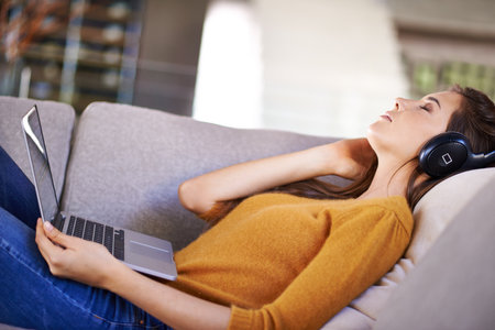 Home, relax and woman with headphones, laptop and streaming music with podcast and peace. Apartment, girl on a couch and person with computer and headset with audio and listening to sound and radioの写真素材