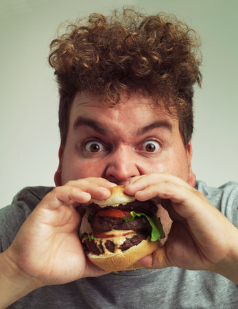 Takeaway, burger and food for man in studio with meal for big eater, plus size or greed. Young person with meat in mouth for hunger, silly and bite indoor for delicious, taste and sauce for portraitの写真素材