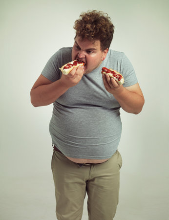 Plus size man, fast food and eating hotdogs for lunch in studio background for snack, hunger and craving. Male person, hungry and takeaway meal with ketchup or starving and enjoy while standingの写真素材