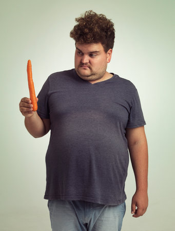 Man, vegetables and carrot in studio with thinking for healthy snack, detox and vitamin c for wellness. Plus size, male person and unhappy with organic food for nutrition, lifestyle and dietの写真素材