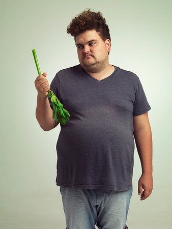 Plus size, diet and unhappy man with vegetable for health, nutrition and wellness in studio in white...