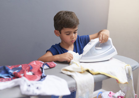 Child, boy and ironing in home with housework for cleaning, sad and punishment with routine or danger. Kid, clothes and housekeeping for responsibility, learning or chores with independence in loungeの写真素材