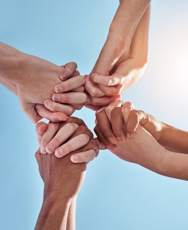 People, team and holding hands together below for unity, collaboration or synergy on a blue sky background. Closeup or low angle of group or community in solidarity for care, support or missionの写真素材