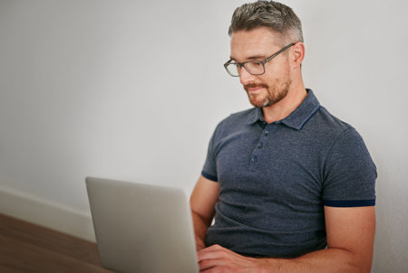 Man, floor and wall on laptop with sitting for relax in casual for social media with technology for internet. Male person, glasses and house with website for blog with typing for freelance writingの写真素材
