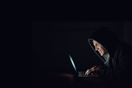 Dark, office and man at laptop for hacking, online programming and cybersecurity at night. Software, developer or hacker in hoodie coding for password, firewall or network access on black backgroundの写真素材