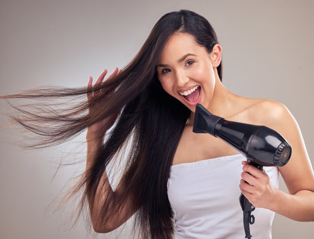 Asian woman, hairdryer and happy with portrait in studio for growth, straighten and brazilian treatment on gray background. Female person, excited and proud with clean hair, keratin and hairstyleの写真素材