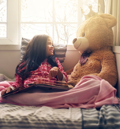 Bed, girl and smile at teddy bear with book in home for storytelling, child development or fun. Happy, literature and kid with stuffed animal in house for fairytale, education or enjoyment in morningの写真素材