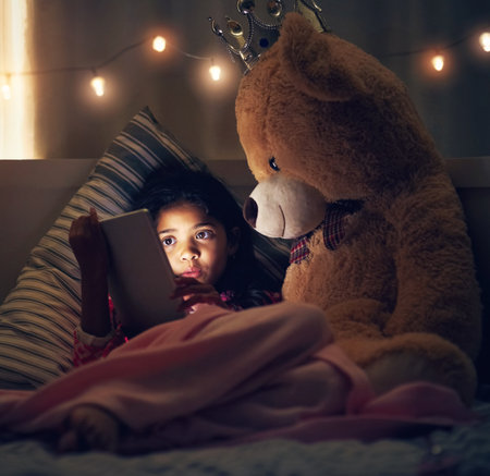 Night, teddy bear and girl with tablet, kid and relaxing with screen light, online reading and social media. Bedroom, home and child with tech, stuffed animal and dark with internet and digital appの写真素材