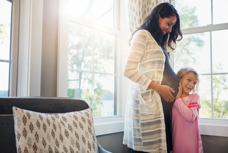 Pregnant, mother and daughter with listening to stomach for baby kick, excited for newborn and curious with smile in home. Woman, pregnancy and happy girl with ear on tummy for bonding, care and loveの写真素材