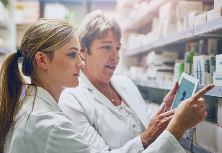Pharmacist, women and together with tablet by shelf for stock, check and inventory with pointing in shop. People, mentor and intern with digital touchscreen, app and inspection for drugs at storeの写真素材