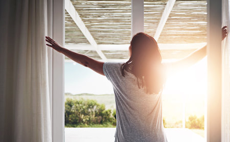 Back, woman and open curtain in morning, window and fresh wake up with sunrise in hotel ready for day. Routine, outside view and nature on holiday or weekend, drapes and light in apartment in bedroomの写真素材