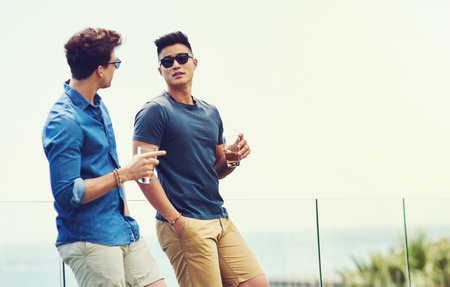 Man, friends and conversation with drinks outdoor on summer vacation for memories, chill and socializing for fun. Male people, glasses and alcohol for enjoyment, speaking and holiday in Santorini.の写真素材