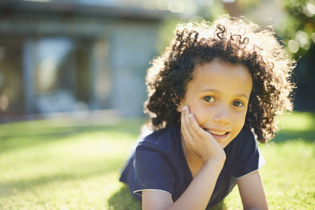 Smile, nature and portrait of child in backyard relaxing on grass for development or fun in sunshine. Happy, sweet and cute boy kid laying on lawn in outdoor garden with positive attitude at home.の写真素材