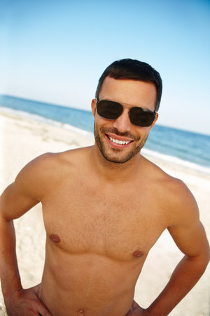 Portrait, happy man, and sunglasses at Brazilian island with blue sky for summer vacation, topless and skin. Smile, beach and face of male tourist with water in outdoor for travel, sunny and holidayの写真素材
