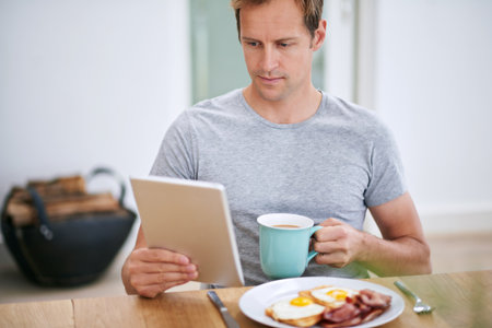 Man, tablet and morning breakfast in home or nutrition meal with coffee, eggs or reading. Male person, relax and digital news in apartment kitchen with food or online article, streaming or connectionの写真素材