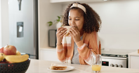 Child, sandwich and eating lunch in home kitchen or healthy nutrition with orange juice, student or breakfast. Girl, kid and morning hungry or bread snack in apartment for fiber meal, diet or foodの写真素材