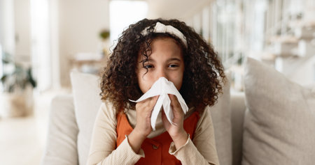 Sick girl, sofa and blowing nose with tissue for virus, infection or congestion in living room at home. Young female person, child or kid with cold, flu or sinus in fatigue, fever or sneeze at houseの写真素材