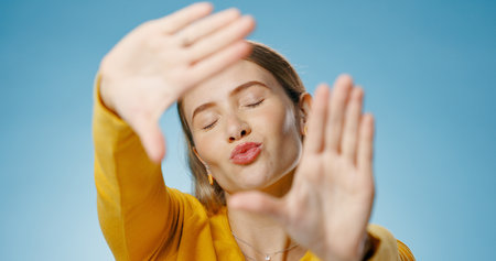 Woman, finger and frame with happy kiss or emoji face, photographer perspective for social media post or picture. Blue background, hand gesture and cosmetic beauty, glow and confident for captureの写真素材