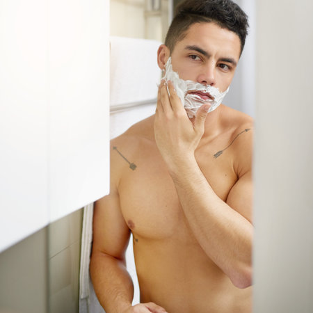 Man, shaving cream and mirror in home bathroom for hygiene, cleaning and check face in morning. Person, cosmetics and topless in reflection, grooming and wellness with foam, change and self careの写真素材