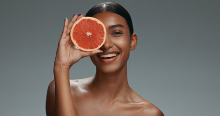 Woman, face and skincare with grapefruit, studio or shine with health, wellness or beauty by dark background. Girl, model and cosmetics with fruit, nutrition or glow with facial aesthetic in portraitの写真素材