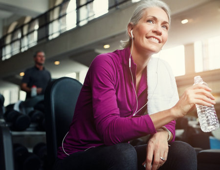 Fitness, relax and senior woman in gym with water bottle for training, exercise and workout. Sports, retirement and person listening to music, resting and drinking for wellness, health and hydrationの写真素材