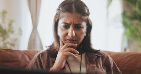 Thinking, woman and laptop on sofa for confused, concerned or internet browsing in home. Female person, thoughtful or deep frown with technology on couch for streaming news or reading blog postの写真素材
