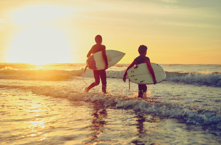Sunset, beach and children with board to surf on adventure in summer vacation and holiday. Kids, learning and swimming in sea on island with waves to exercise with safety in nature or energy outdoorの写真素材