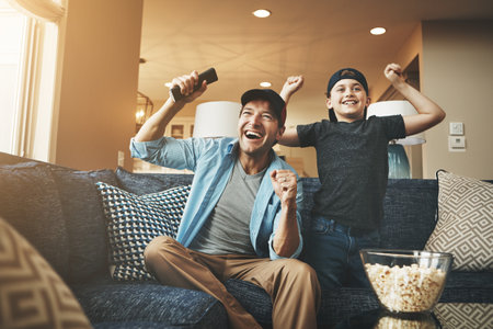 Television, popcorn and man with kid in celebration for broadcast, subscription and bonding at home. Father, son and happy with tv remote in fist for sports, success or watching live network togetherの写真素材