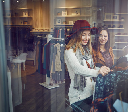 Shopping, happy and girl friends by clothes in retail mall for discount, sale or promotion. Style, window and young women bonding and buy designer outfit on rack at luxury fashion boutique or store.の写真素材