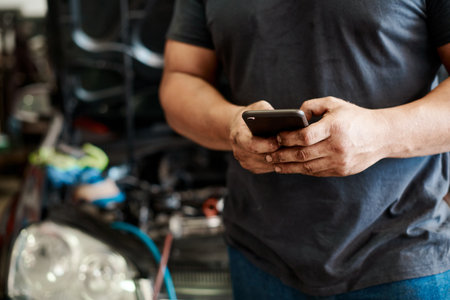 Phone, hands and mechanic in workshop with car service, maintenance or search for auto insurance. Typing, engine and man in garage with smartphone for inspection, motor repair and quality assuranceの写真素材