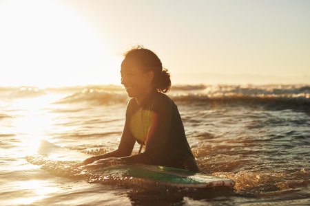 Surfer, woman and swimming in ocean with wetsuit for summer adventure, sunset and fun activity in water. Girl, surfing sport and floating in waves with board for vacation travel, exercise and peaceの写真素材