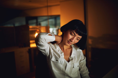 Asian woman, neck pain and tired in night at office with stress, injury or muscle fatigue at startup. Person, frustrated or burnout with massage, joint or exhausted at creative agency in South Koreaの写真素材