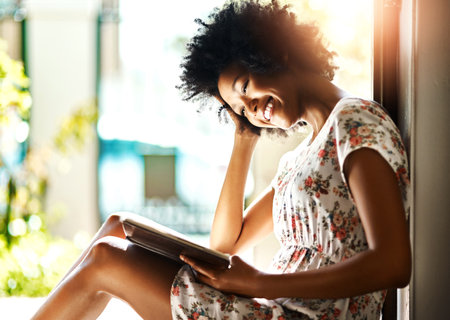 African woman, reading book and smile by window, sunshine and relax with poetry in morning at apartment. Person, happy and literature for knowledge, learning and story in fantasy novel in houseの写真素材