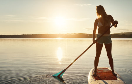 Lake, sunset and woman on paddle board for balance, water sport and vacation on river for tourism. Nature, summer or holiday in Germany for female person, swimsuit or travel exercise for adventureの写真素材