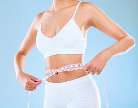 Woman, measuring tape and stomach in studio with diet for weight loss with body in underwear for health. Slim, waist and detox with abdomen isolated on blue background with fitness, skin and wellnessの写真素材
