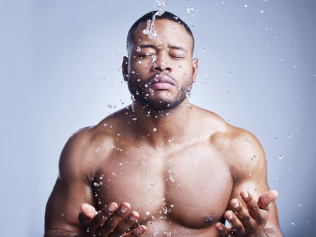 Black man, beauty and water drop for washing face in studio, cleaning and hydration on blue background. Male person, liquid and skincare for facial treatment, fresh and cosmetics for grooming routineの写真素材