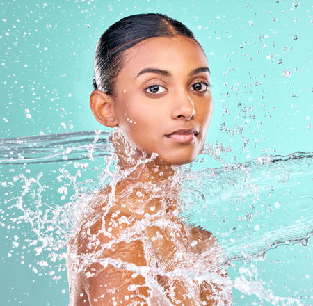 Indian woman, portrait and water splash for skin care, hygiene and beauty or dermatology in studio. Face of model with liquid for waterproof cosmetics, makeup or facial washing on blue backgroundの写真素材