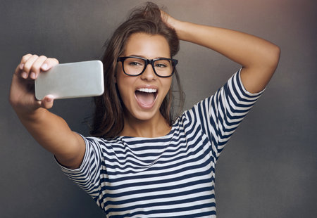 Selfie, smile and woman with glasses, picture and happiness with social media on grey studio background. Person, human and model with smartphone, connection and memory with reaction and expressionの写真素材
