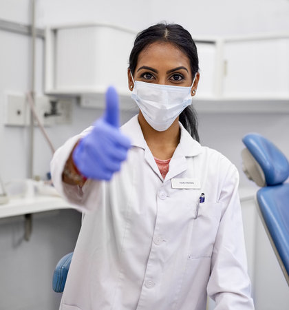 Woman, thumbs up and mask face in dentist, dental health and covid protection with hygiene professional in practice. Oral care, gloves and safety with orthodontics employee, agree and hand gestureの写真素材