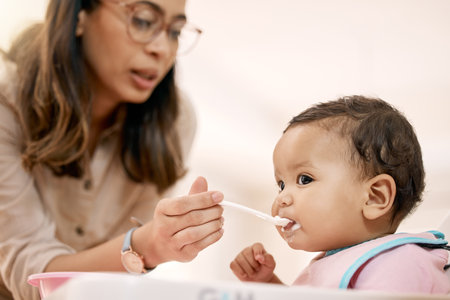 Woman, baby and spoon feeding for nutrition, growth and child development in home for diet or motor skills. Infant, mother and eating with hungry, care and love for health, vitamins and milestoneの写真素材