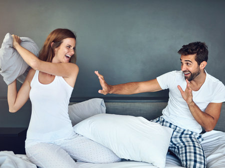Love, pillow fight and romance with couple in bed together for bonding, excitement or having fun. Happy, playful or smile with man and woman in bedroom of home for morning comedy or silly gameの写真素材