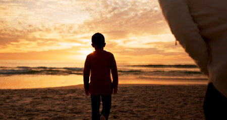 Boy, mother and walking at sunset on beach, travel and explore nature or waves of ocean on vacation. Kid, mommy watch and sea for wellness or back in Florida on weekend, water and holiday adventureの写真素材