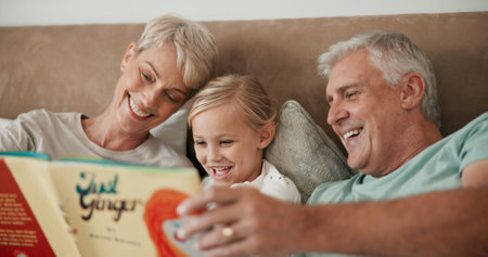 Grandparents, girl and reading book in bed to relax, literature and literacy for bonding at sleepover. Home, grandchild and people learning for love, language and grandma or grandpa for fairytaleの写真素材