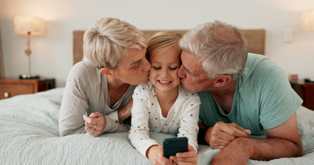 Grandparents, kissing and mobile phone with grandchild in bedroom for growth, development and gaming in home. Girl, happy and tech for learning, joy and fun as youth, kid or young person with bondingの写真素材