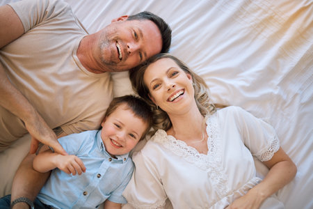 Happy family, portrait and lying with child above on bed for bonding, holiday or weekend at home. Top view of mother, father and son with smile or relax in bedroom for morning, rest or joy at houseの写真素材