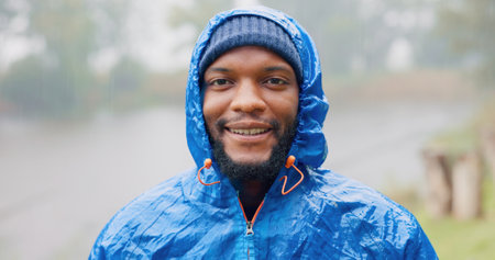 Black man, portrait and rain for travel outdoor on camping adventure, explore and nature journey in morning. African, male person and smile for hiking, freedom and trekking woods for winter vacationの写真素材