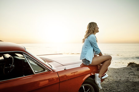 Road trip, woman and adventure with beach, sunset and view for Miami holiday and vacation. Muscle car, hood and thinking with idea, freedom and summer for driving and journey with ocean sightseeingの写真素材